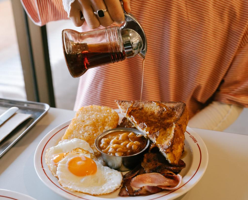 syrup being poured onto a big plate of pancakes, eggs and beans by a women in an orange shirt 