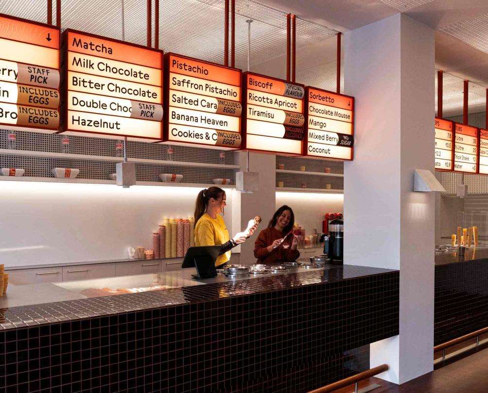 an image of two woman behind the counter at an ice cream shop with flavours of ice cream up above
