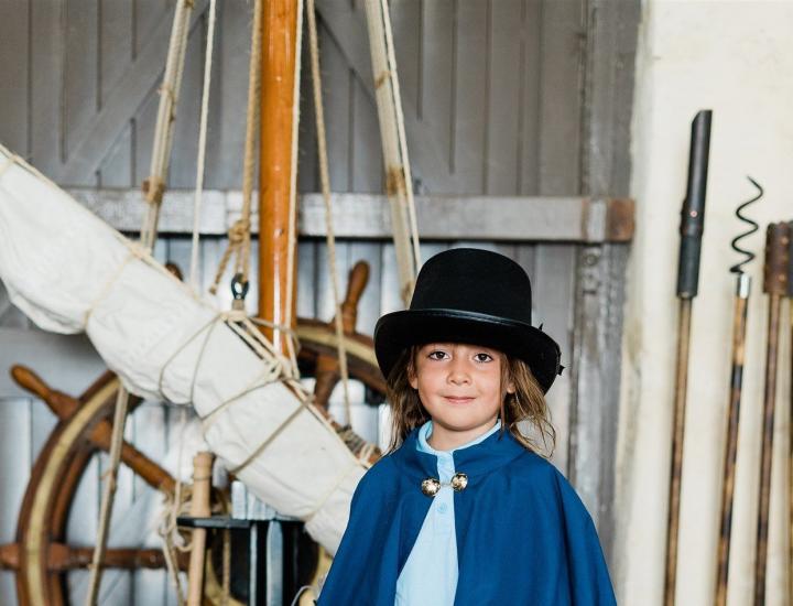 Young boy wearing a black bowler hat and a blue cape with various old-fashioned sailing equipment in the background.