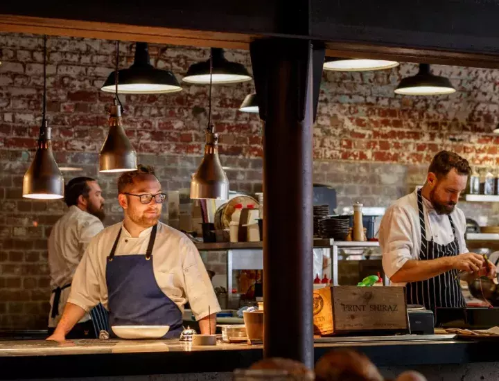 two chefs cook in contemporary restaurant kitchen surrounded by fresh produce. smiling person walks past