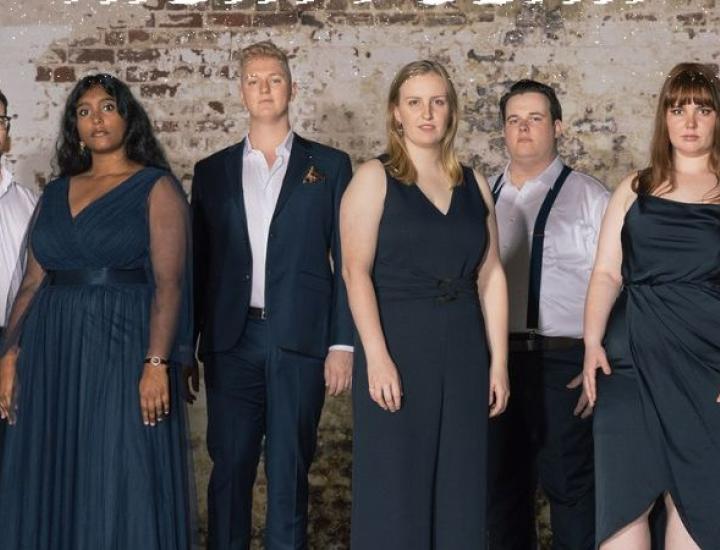 ten performers dressed in navy and white stand against an exposed brick wall