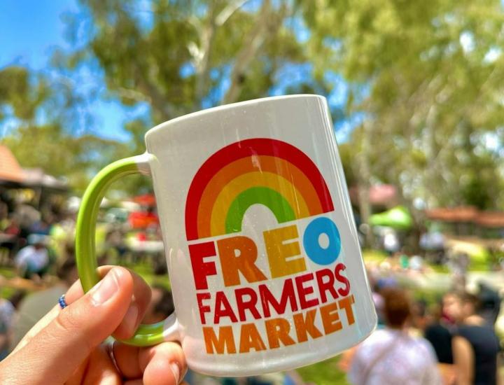 hand holds mug against blue sky and busy outdoor markets. mug shows rainbow and the words 'FREO FARMERS MARKET'