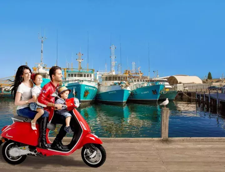 Family driving a Vespa by Fishing Boat Harbour in Fremantle with blue fishing boats in the background