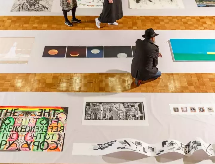A man kneels on a floor surrounded by various artworks.