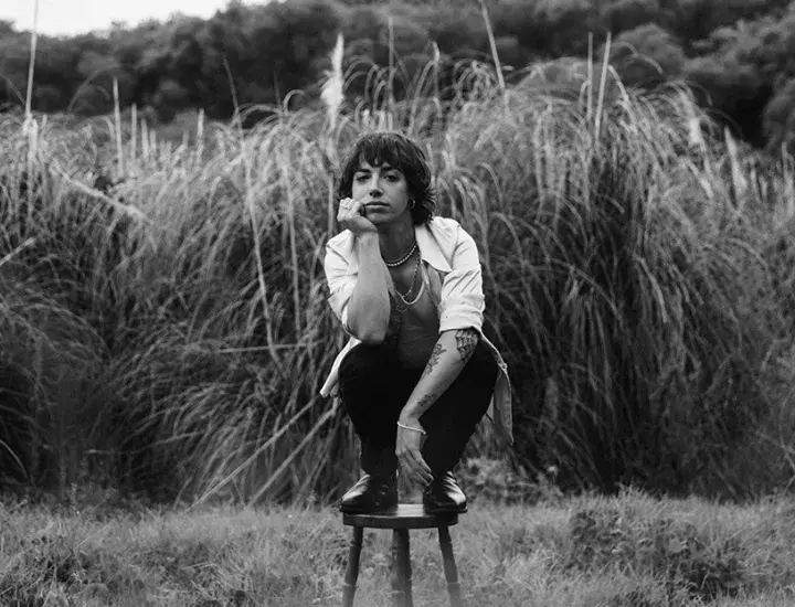 A man with dark hair in a white shirt and black pants poses on a stool in front of a field.