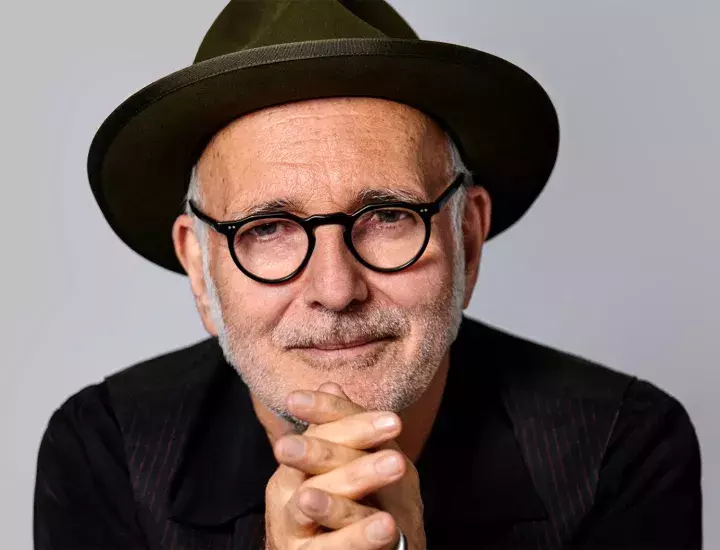 A close up of a man in black spectacles and hat with his hands held together under his chin.