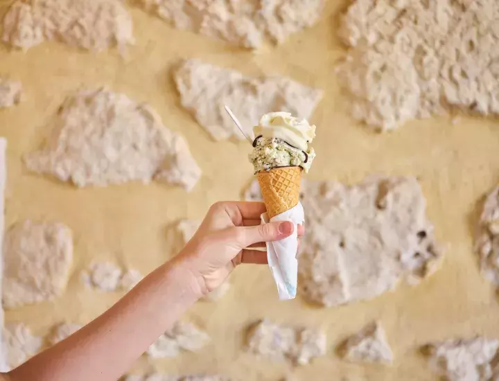A hand holding an icecream cone with a scoop of pistachio flavoured icecream on top.