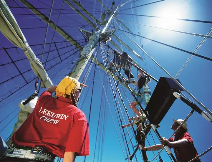 A crew member of a tall ship watches as three other crew members climb a mast.