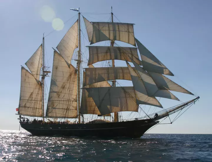A tall ship with several masts sails on the ocean under a blue sky.